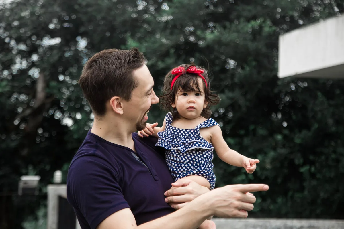 A parent carrying a young child in a backyard, one hand braced on their lower back.