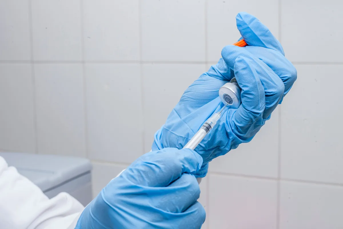 Gloved hands drawing medication from a small vial into a syringe.