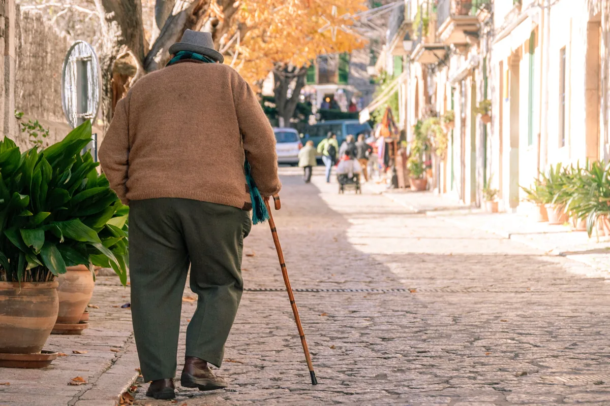 An older person seen from behind, walking slowly down a street with a cane.