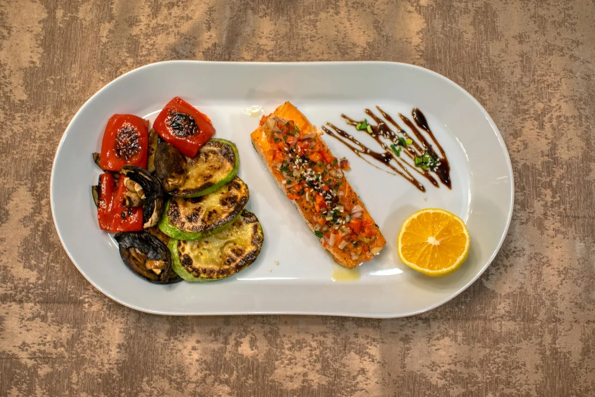 Grilled salmon fillet beside grilled zucchini, eggplant and red pepper, with half an orange, on a white oval plate.
